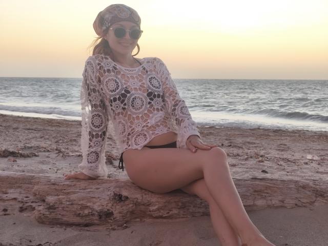 SarahRousen with a white lace top, black bikini bottom, sunglasses, and headscarf, sitting on a driftwood log at sunset, showing her ass and legs.