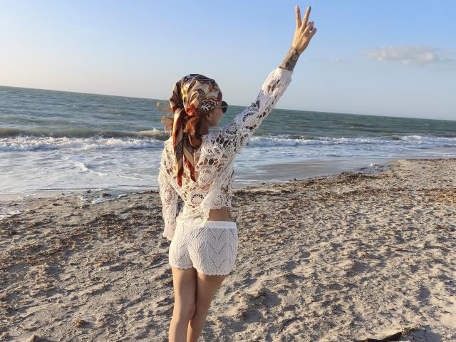 SarahRousen standing on a beach, wearing a white crocheted top and shorts, raising her right arm, long curly hair with red and brown accents, ocean in background.