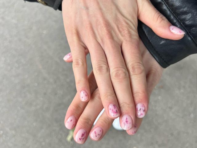Le mani di Valnes, con le dita delicatamente sovrapposte, mostrano uno smalto rosa con venature scure. Indossa una giacca di pelle nera e la sua pelle è chiara. Le sue unghie sono curate e ben tagliate..