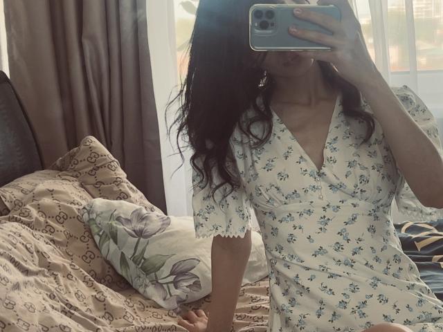 Valnes with wavy dark hair, wearing a white V-neck dress with blue floral patterns, taking a mirror selfie, showing her tits and holding a blue smartphone.