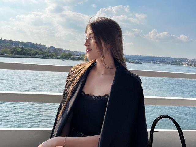 DremaEspina, avec de longs cheveux bruns, un haut noir bordé de dentelle et une étole noire, assise près de l'eau, les yeux fermés, regardant sur le côté, ensoleillement, ciel clair, eau calme, sac noir..