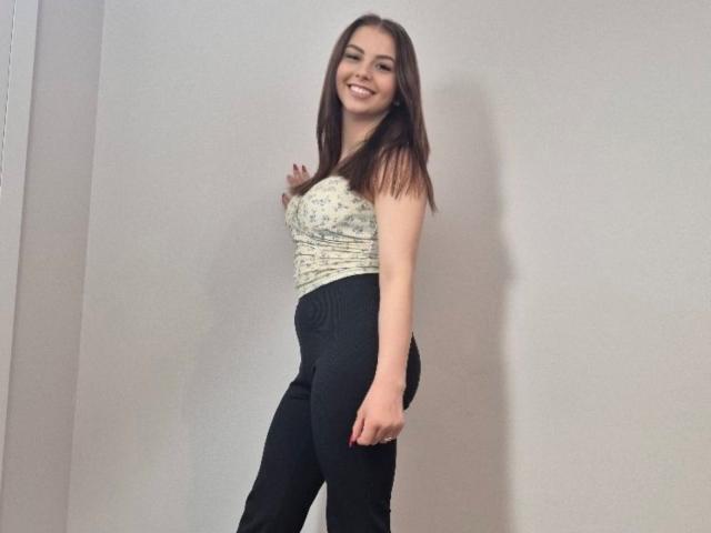 MillyTop tem cabelos castanhos longos, um sorriso, um top branco com padrão e calças pretas de cintura alta, está de lado com a mão na cintura, mostrando o bumbum..
