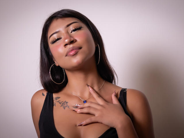BonnieBodn with dark hair, tan skin, hoop earrings, black top, tattoos on right shoulder, hand on chest, blue ring, pink lips, subtle smile, eyelashes, and nose ring.