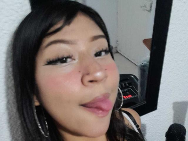 BonnieBodn has dark hair, thick eyelashes, and is sticking out her tongue with pink lips. She's wearing large hoop earrings and has a small mole on her left cheek.