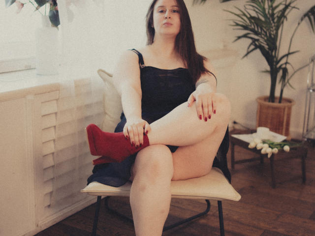 ViaLattea está sentada em uma cadeira, vestindo um vestido preto, saltos altos vermelhos e esmalte vermelho, com uma perna cruzada, expondo suas coxas pálidas e grossas e o pequeno bumbum..