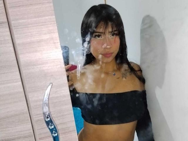 BonnieBodn with long black hair, wearing a black off-shoulder top, showing small tits and a tattoo on her chest, standing in front of a mirror with fog.