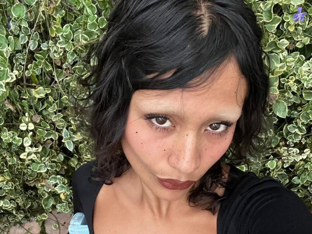 AliceFirek with dark curly hair, brown eyes, and freckles, puckering her brown lipstick-covered lips, wearing a black top, surrounded by green and white leafy plants.