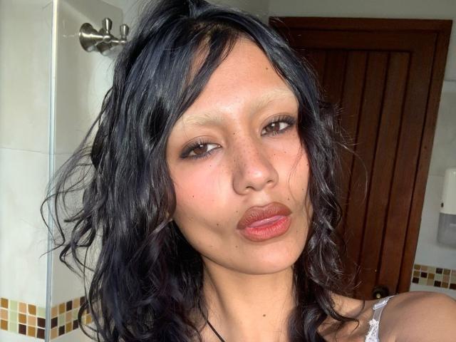 AliceFirek's pouty face with dark wavy hair, brown eyes, and slightly freckled skin, making a cute, puckered-up expression. Her lips are full and her nose is small.