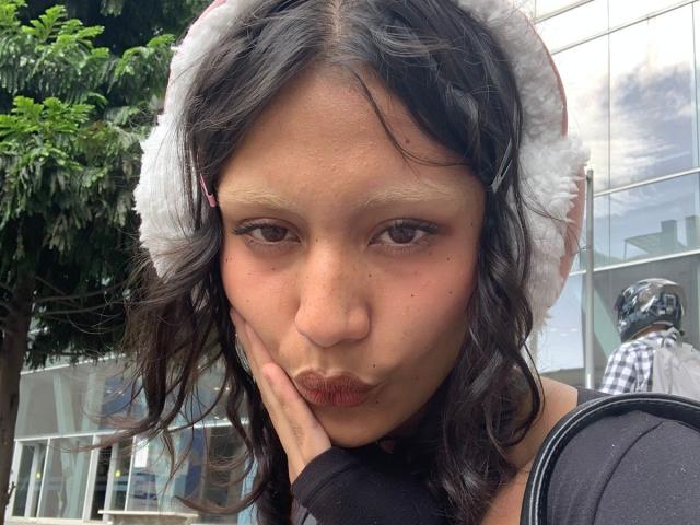 AliceFirek with dark wavy hair, wearing a white fuzzy hat, pouting her lips and resting her hand on her cheek. She's outside with a tree and building in the background.