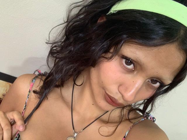 AliceFirek with wavy black hair, green headband, brown eyes, and small nose, pulling down floral strap, showing collarbone, wearing necklace, and slightly smiling.