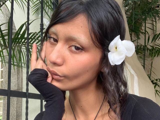 AliceFirek with brown eyes, black hair, and a white flower in her hair, wearing a black long-sleeve shirt, resting her cheek on her hand.