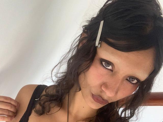 AliceFirek with curly brown hair, black eyeliner, and a white hairpin, wearing a black top, looking up with pouty lips and slightly flushed cheeks.