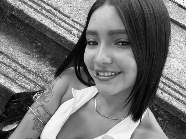 Black-and-white selfie of SaritaKlart with straight dark hair, smiling, wearing a white top, showing a tattoo on her right arm, and a metal bracelet, with hoop earrings.