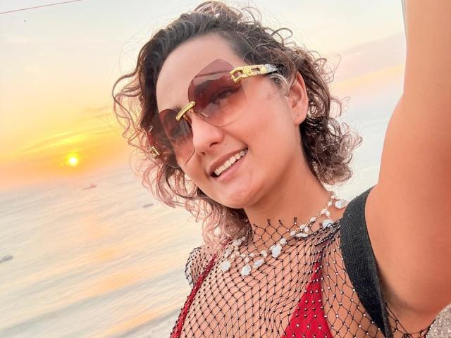 Flora Saenz, avec ses cheveux bruns bouclés, souriante, portant un haut en maille rouge, des lunettes de soleil dorées et un collier de perles blanches, montrant sa poitrine, au coucher du soleil. Son bras est levé pour prendre un selfie..