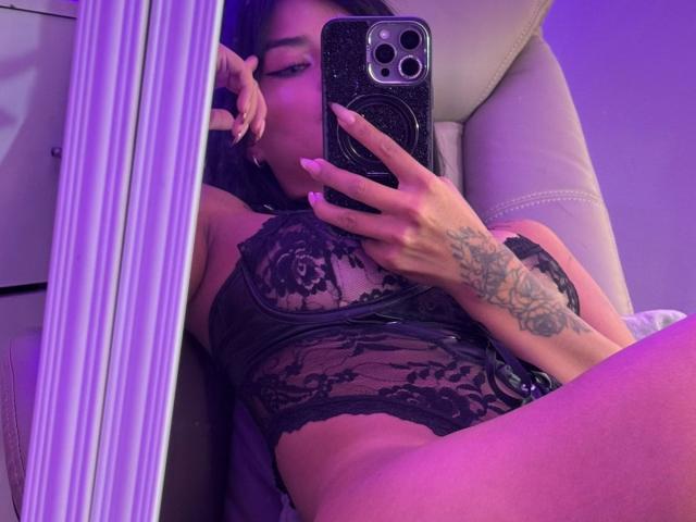 MajoJones in black lace lingerie, holding phone, tattoos on hand, purple lighting, mirror selfie, visible tits, thigh in foreground, relaxed in chair.
