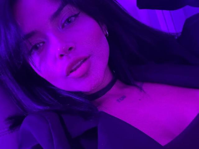MajoJones with purple-lit face, black choker, and black top, showing small tattoo on chest, with long dark hair and subtle makeup, looking at camera with slight smile.