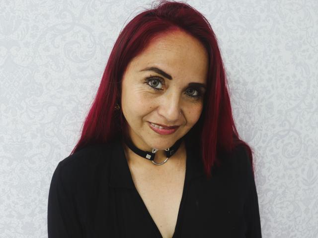 CamilaLux has red hair, green eyes, and a black choker with a ring. She's wearing a black top, smiling subtly, and has small tits. Her skin is light.