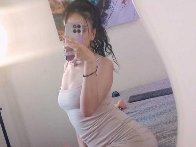 SharonKm indossa un abito bianco aderente con pieghe, che mette in mostra il suo seno piccolo e i suoi fianchi formosi, e sta scattando un selfie allo specchio con il suo telefono. Ha i capelli ricci e scuri, e sullo sfondo si vede un muro bianco..