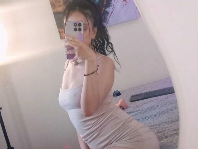 SharonKm, avec ses cheveux noirs ondulés, porte une robe beige moulante et transparente, qui révèle ses petites seins et ses courbes généreuses. Elle prend un selfie devant un miroir avec un téléphone violet..