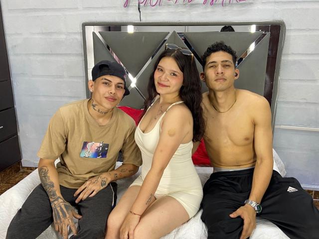 Three PlayMatesX sitting on a bed: um com uma camisa bege e tatuagens, uma com um vestido branco e seios pequenos, e um sem camisa, com calças pretas e um relógio..