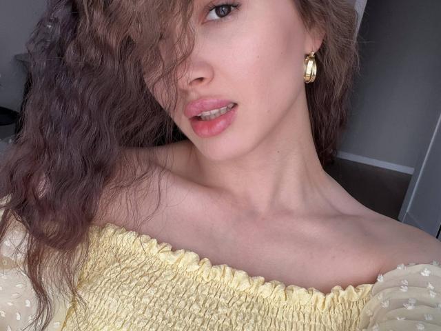 Paulita Grantz mit welligem, braunem Haar, rosafarbenen Lippen und goldenen Ohrringen, trägt ein gelbes Top mit einem Schulterfrei-Schnitt, das ihren nackten Hals und das obere Brustteil zeigt. Brüste sind nicht sichtbar..