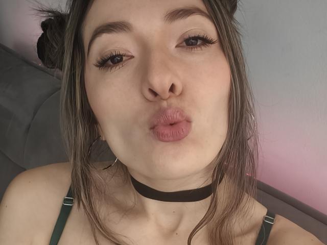 TaniaSandersX tiene labios rosados y ligeramente fruncidos, un collar grueso negro, tirantes verdes y cabello castaño recogido en moños despeinados. Lleva aros grandes, delineador de ojos discreto y pestañas largas..