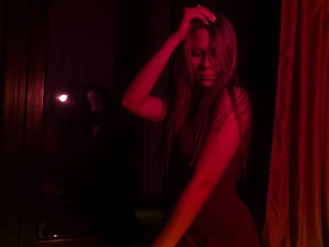 MichelinaConeway with long blonde hair, red-lit, wearing a dark dress, hand in hair, sensual, looking down, dark room, red curtain on the right, shadowy figure in background.