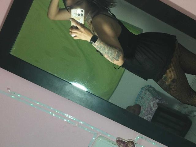 TaylorLees with tattoos, black sleeveless top, black mini skirt, and fishnet stockings, taking a mirror selfie showing her ass, in a pink room with a green wall.