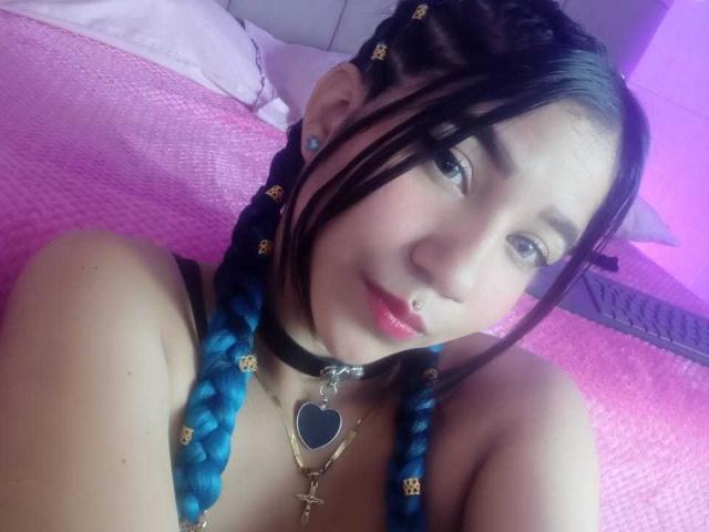 TaylorLees has blue braids, a black choker with a heart pendant, a nose ring, and a cute, slightly shy expression. Her pink bed is in the background.