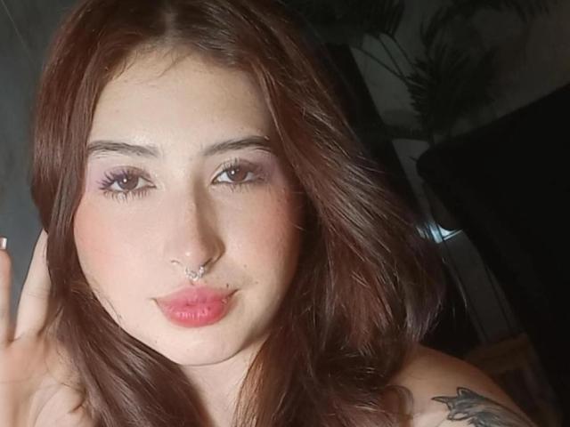 Mia Volkova, avec ses cheveux roux, ses paupières roses, un anneau au nez, ses lèvres roses et un tatouage sur son épaule droite, faisant une moue et tenant ses cheveux avec sa main gauche..