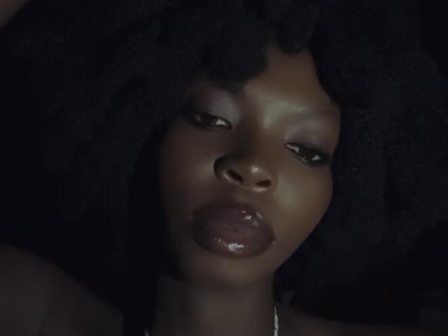Le labbra marrone scuro, gli occhi marrone scuro e i capelli ricci neri e lucidi di DarkMajesty sono al centro dell'attenzione. La sua pelle scura e liscia contrasta con lo sfondo scuro..