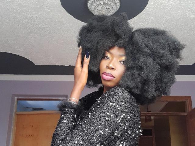 DarkMajesty, com um grande cabelo afro preto, vestindo um vestido preto brilhante, tem batom rosa e esmalte de unha escuro, e está tocando no cabelo. Luz no teto acima..