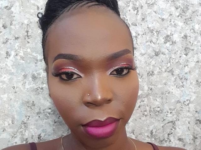 DarkMajesty has shiny brown skin, pink lipstick, thick black eyebrows, long eyelashes, and braided hair. She's wearing a silver necklace against a textured gray wall.