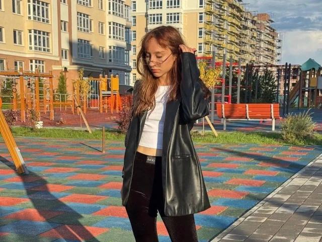 ModelGyrl: bella, con lunghi capelli castani, top bianco, giacca di pelle nera e pantaloni neri, in piedi in un colorato parco giochi, con le mani tra i capelli, la luce del sole sul viso..