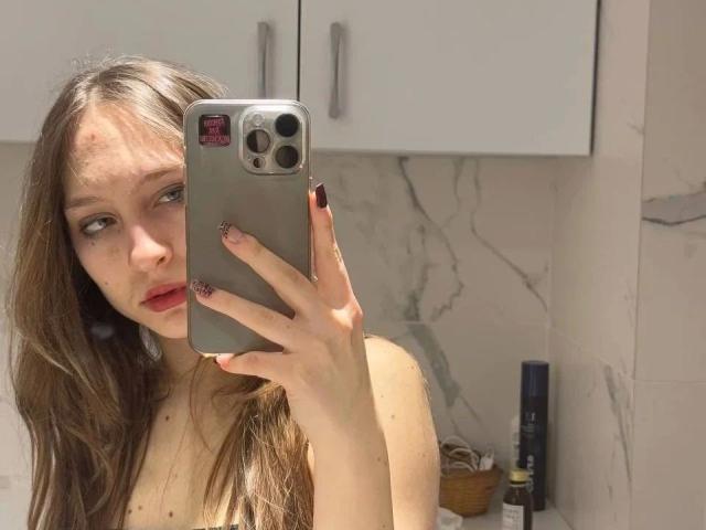 Modelo atractiva con cabello castaño largo, lápiz labial rojo y piel clara, sosteniendo un iPhone plateado, mostrando sus pechos desnudos, de pie en un baño con paredes de mármol..