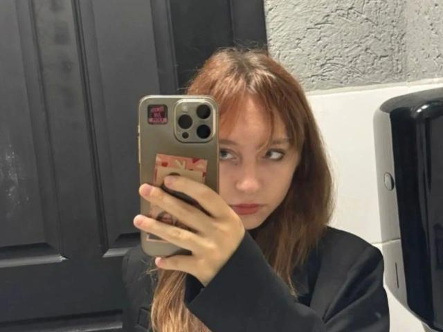 Modelo atractiva con cabello rojo, tomando una selfie, sosteniendo un teléfono con una funda bonita, vistiendo una blusa negra, con una expresión ligeramente molesta, en un baño..