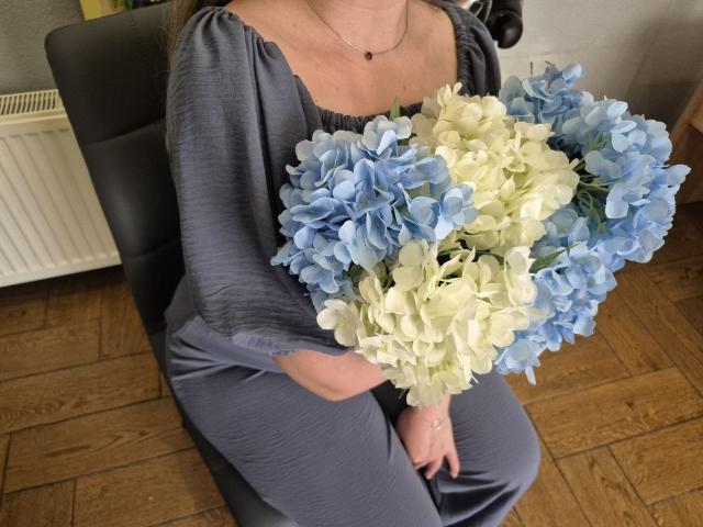 Meeranda con il seno scoperto, indossa un vestito grigio, tiene un bouquet di ortensie blu e bianche, è seduta su una sedia nera, pavimento in legno, termosifone sullo sfondo..