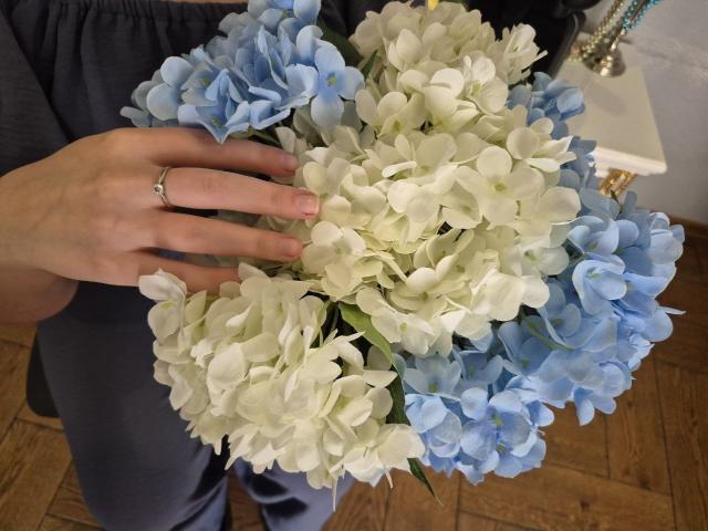 Meeranda's hand holds a bouquet of white and blue hydrangeas. Her finger with a simple ring rests on the flowers. Wooden floor and Meeranda's dark pants visible.