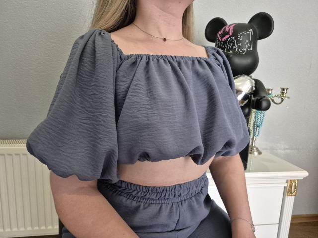 Meeranda, vestindo uma blusa cinza de manga bufante que deixa sua cintura à mostra, e calças cinzas combinando, sentada em frente a uma estatueta preta do Mickey Mouse com um laço rosa..