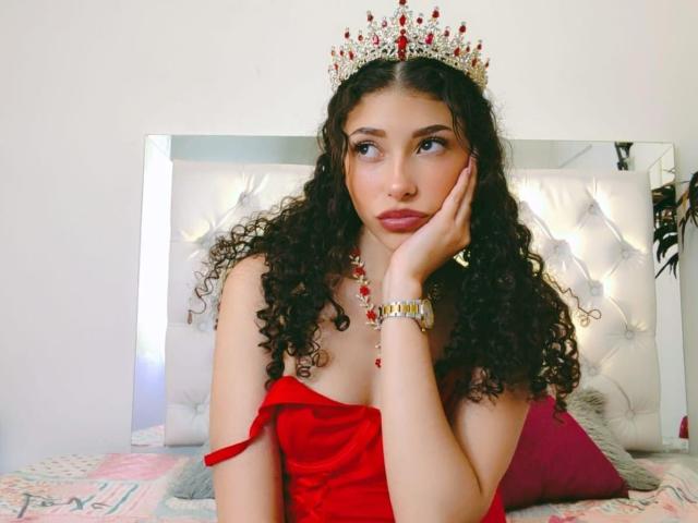 SophiaLuthor with curly black hair, wearing a red off-shoulder dress, jeweled crown, and red necklace, resting her face on her hand, looking to the side.