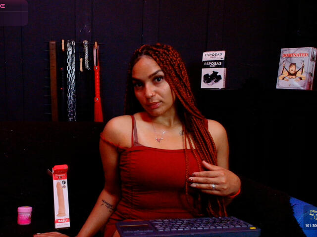 BrianaPratt with red braids, wearing a red dress, sitting at a computer, holding her right hand near her chest, showing her tits, in a dimly lit room.