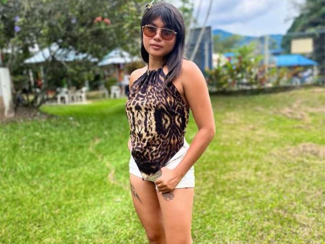 AshlyLee with tan skin, black hair, and sunglasses, wearing a leopard print halter top and white shorts, showing a tattoo on her right thigh, standing on green grass.
