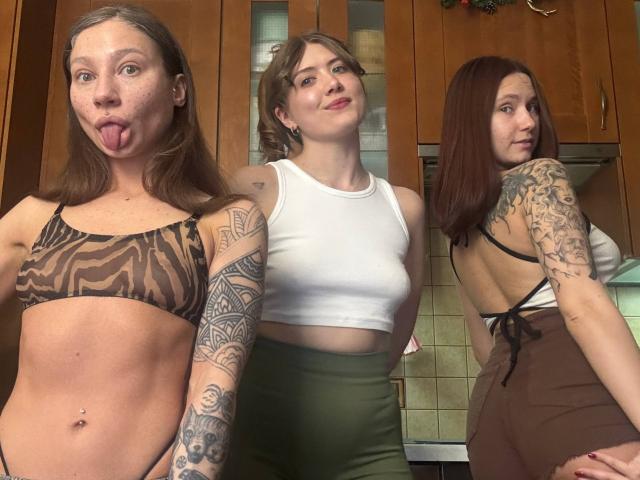 Three HotCaramelsTrio with tattoos, wearing zebra bikini, white crop top, and brown apron, sticking tongues out, in a kitchen. One has a belly button piercing.