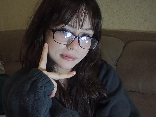EileneSzala with dark brown hair, glasses, and a green fleece, touching her cheek with a finger, smiling softly. Her ass is not visible.