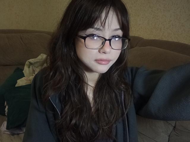 EileneSzala with long brown hair, glasses, and pink lips, wearing a black zip-up, holding out her arm, sitting on a brown couch with a green pillow.