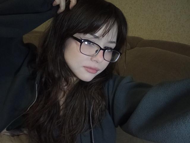 EileneSzala with black glasses, long wavy brown hair, pink lips, and a black hoodie, taking a selfie with her right arm raised, showing a calm, slightly sexy expression.