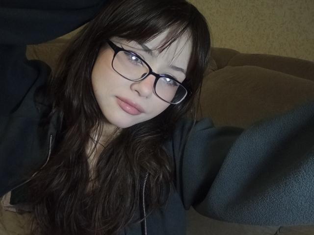 EileneSzala has long brown hair, glasses, and pale skin, wearing a dark hoodie, with a neutral expression, lying on a brown couch, arms raised.
