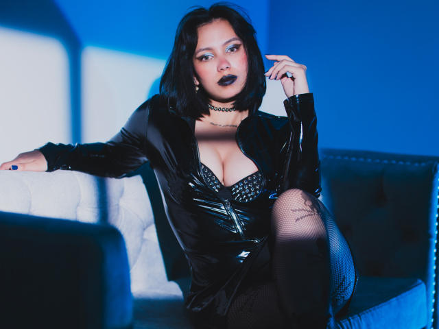 Lilith Deveraux mit einem schwarzen Bob, trägt ein glänzendes schwarzes Latekleid, zeigt ihre Brüste in einem mit Nieten besetzten BH, trägt Netzstrümpfe und eine schwarze Halskette, und sitzt auf einem blau beleuchteten Sofa..