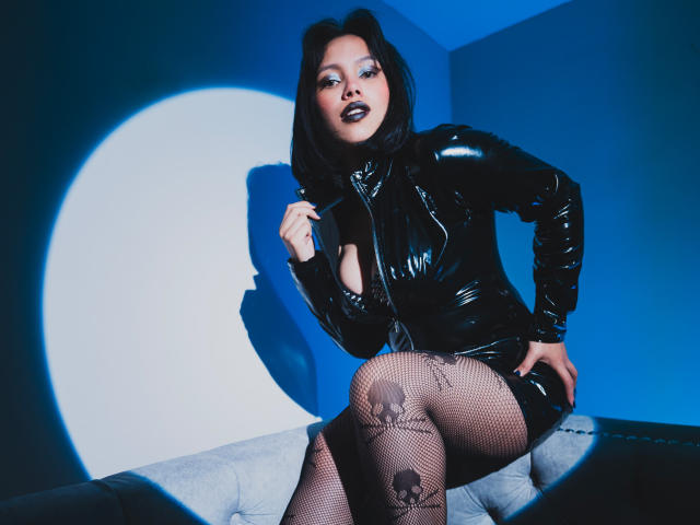 LilithDeveraux vestindo um vestido de látex preto brilhante, meias de rede pretas com estampa de morcego, sentada em um sofá cinza, com a mão no peito, e a sombra do rosto dela projetada em uma parede azul..