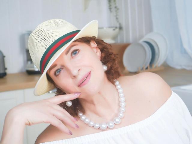 LinaHotMom-sex with curly red hair, blue eyes, wearing a white off-shoulder top, pearl necklace, and straw hat with green and red band, touching her chin with a finger.